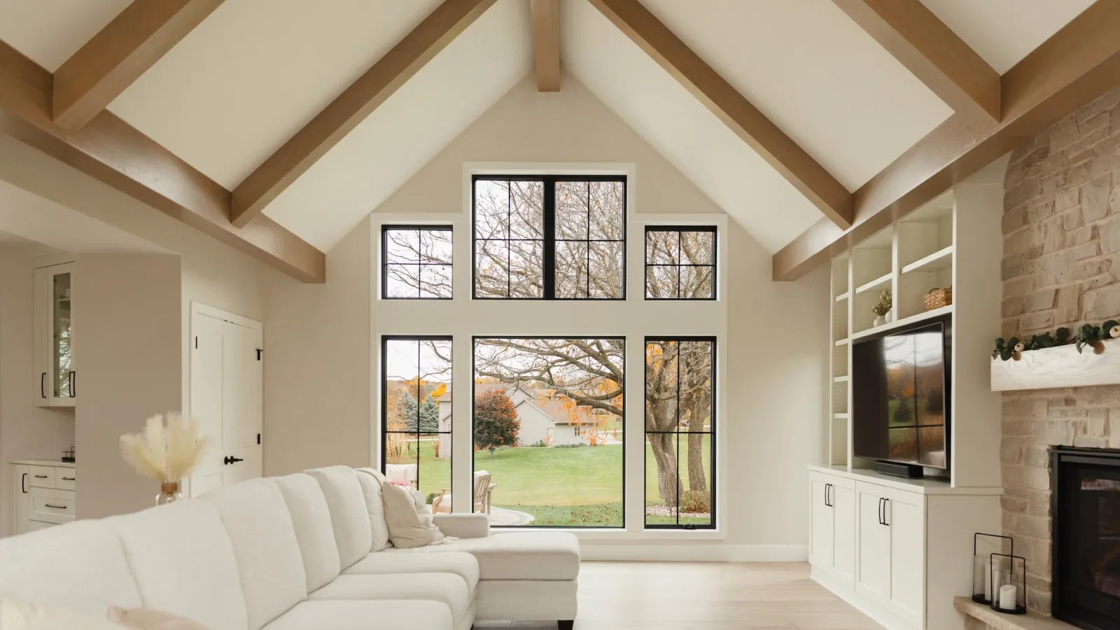Bright living room with vaulted wooden beams, large black-framed windows, white sectional, and stone fireplace.