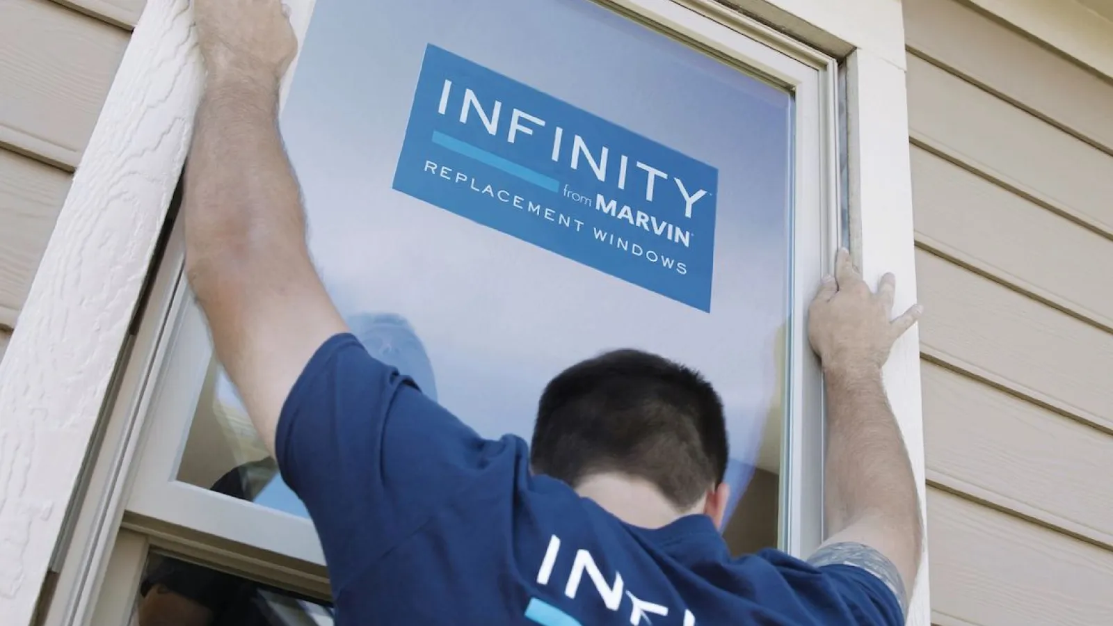 Worker installing an Infinity from Marvin replacement window on a beige house exterior during home renovation.