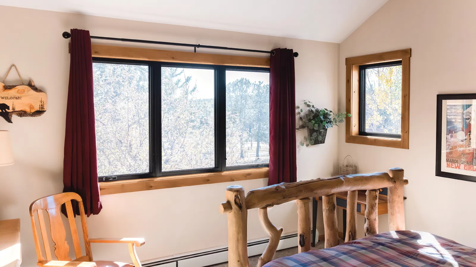 Rustic bedroom with log bed frame, plaid bedding, wooden chair, burgundy curtains, and large window with trees outside.