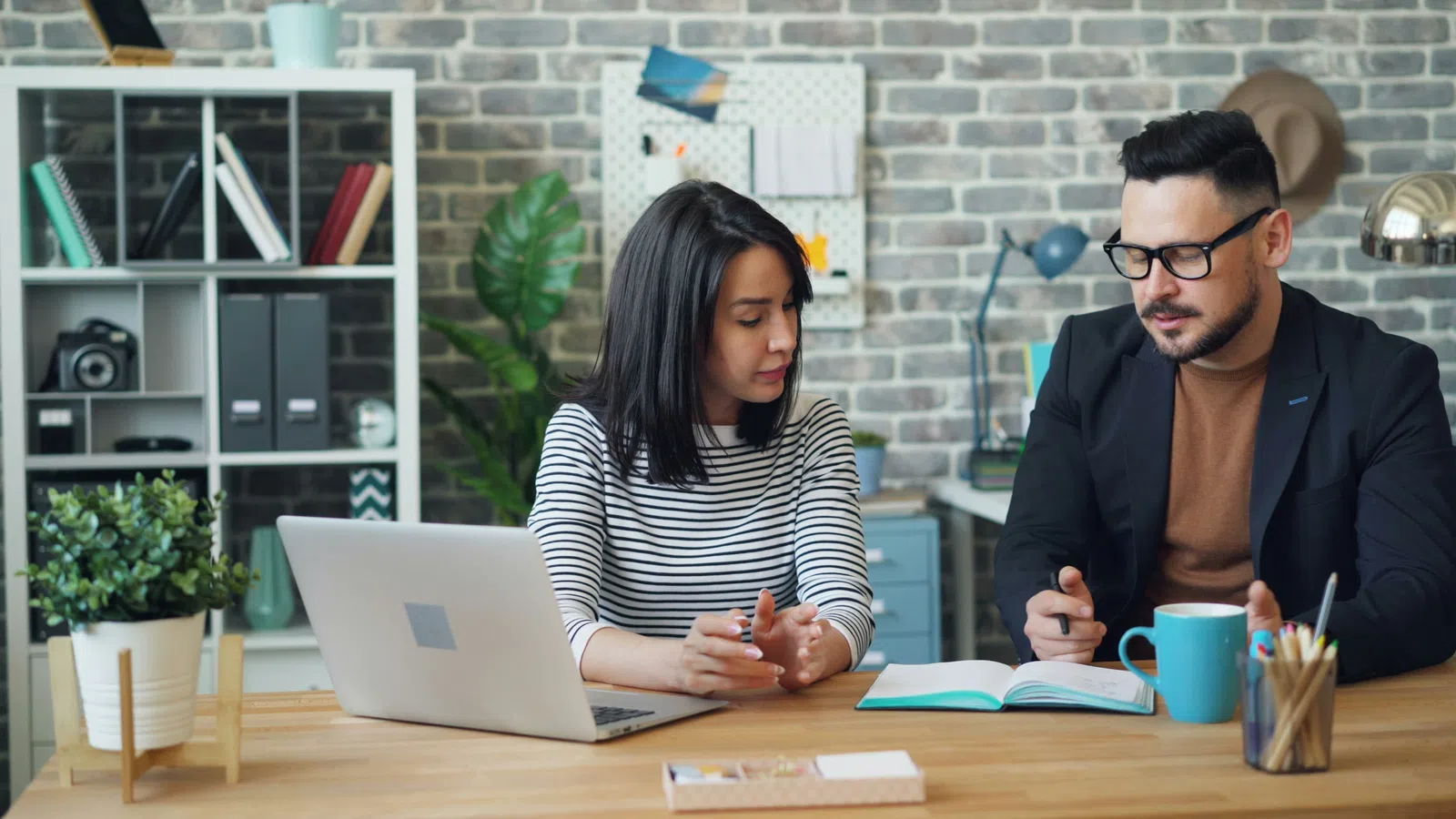 Two colleagues discussing work at a wooden desk with a laptop, notebook, and office supplies in a modern workspace