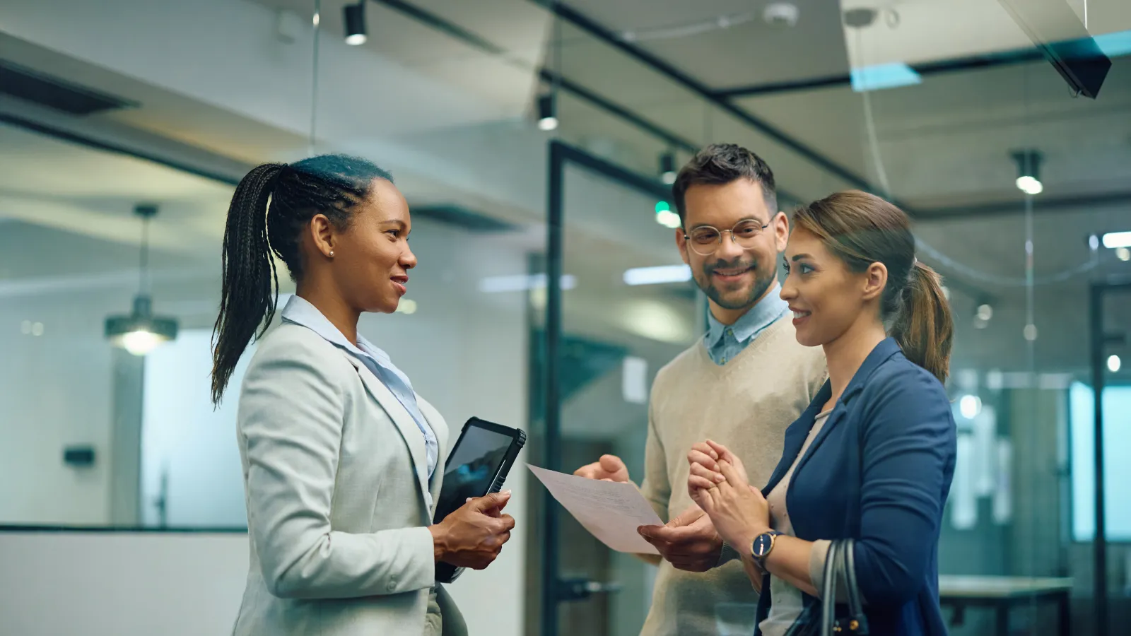 Three professionals engage in a discussion inside a modern office with papers and a tablet.