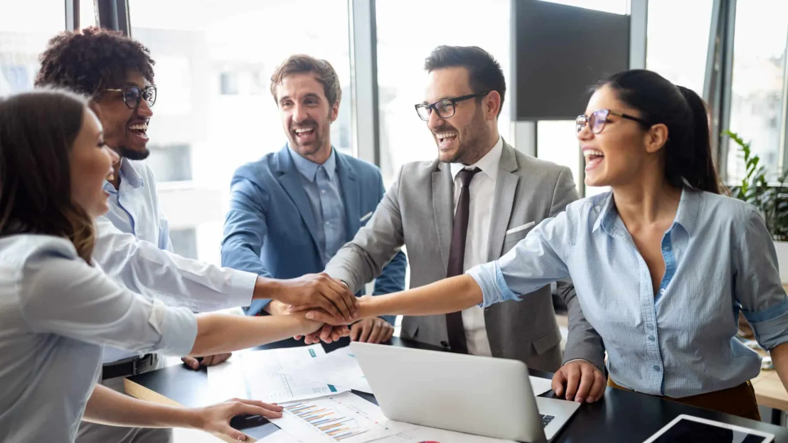 Diverse business team joining hands in office meeting for teamwork and success with charts and laptop