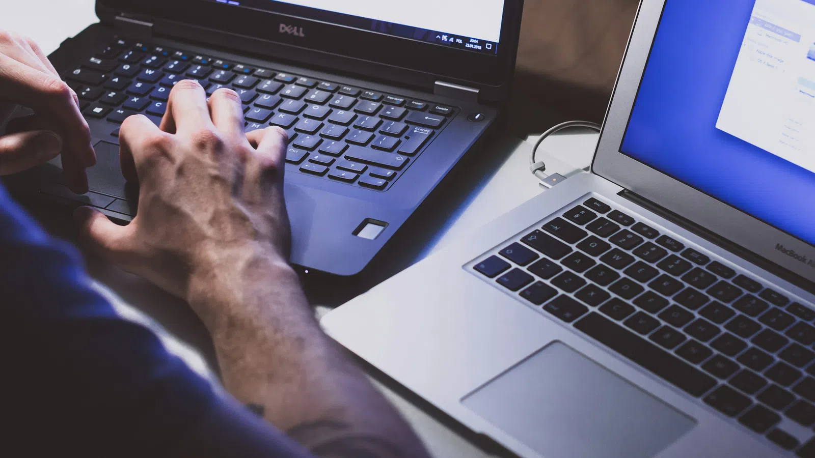 Person typing on Dell laptop next to an open MacBook Air on a desk with screens showing software windows