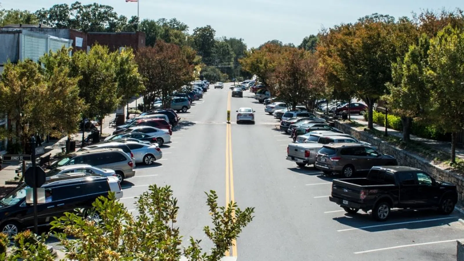 Quiet small-town street lined with parked cars, trees, and buildings under a clear sky midday.