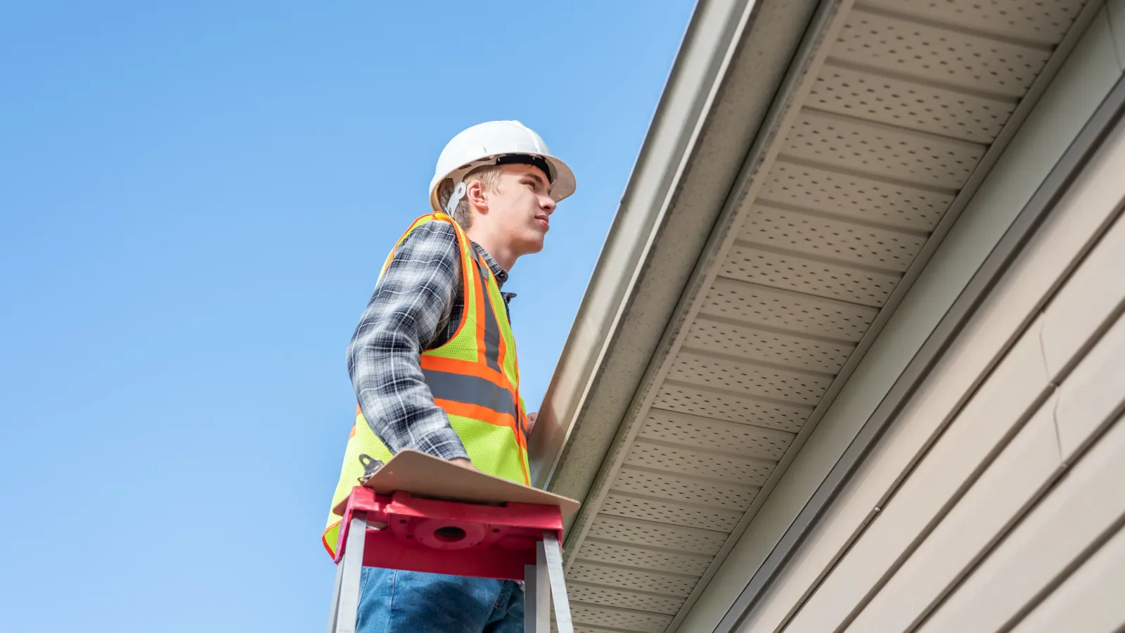 Construction worker in safety gear inspecting house exterior while standing on ladder under clear blue sky