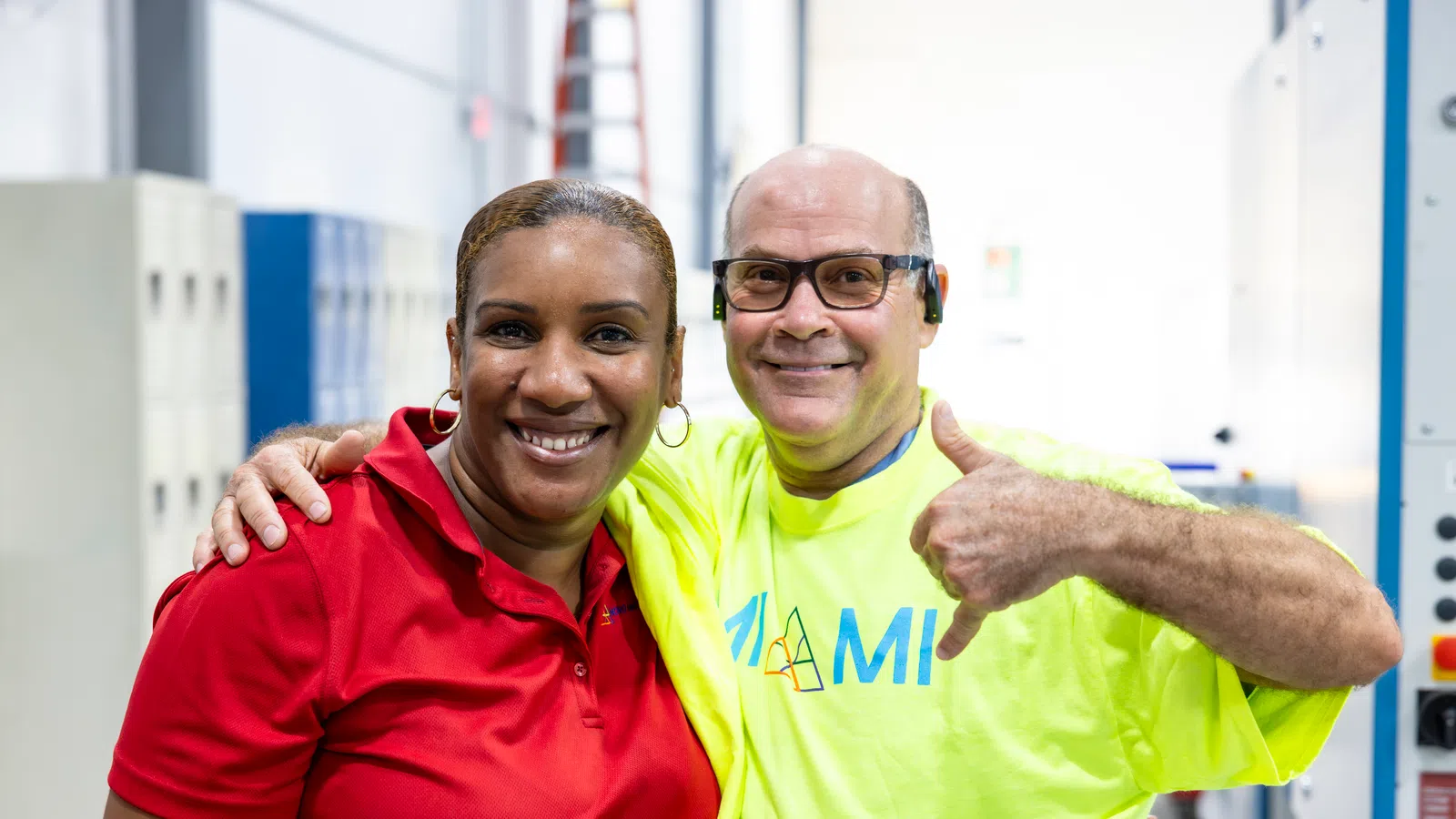 Two factory workers smiling together, one in red shirt and the other in yellow, showing a thumbs-up gesture indoors.