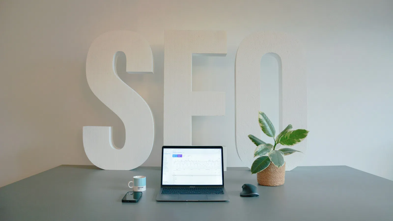 Workspace with laptop showing SEO data, smartphone, coffee mug, plant, and large white SEO letters in background.