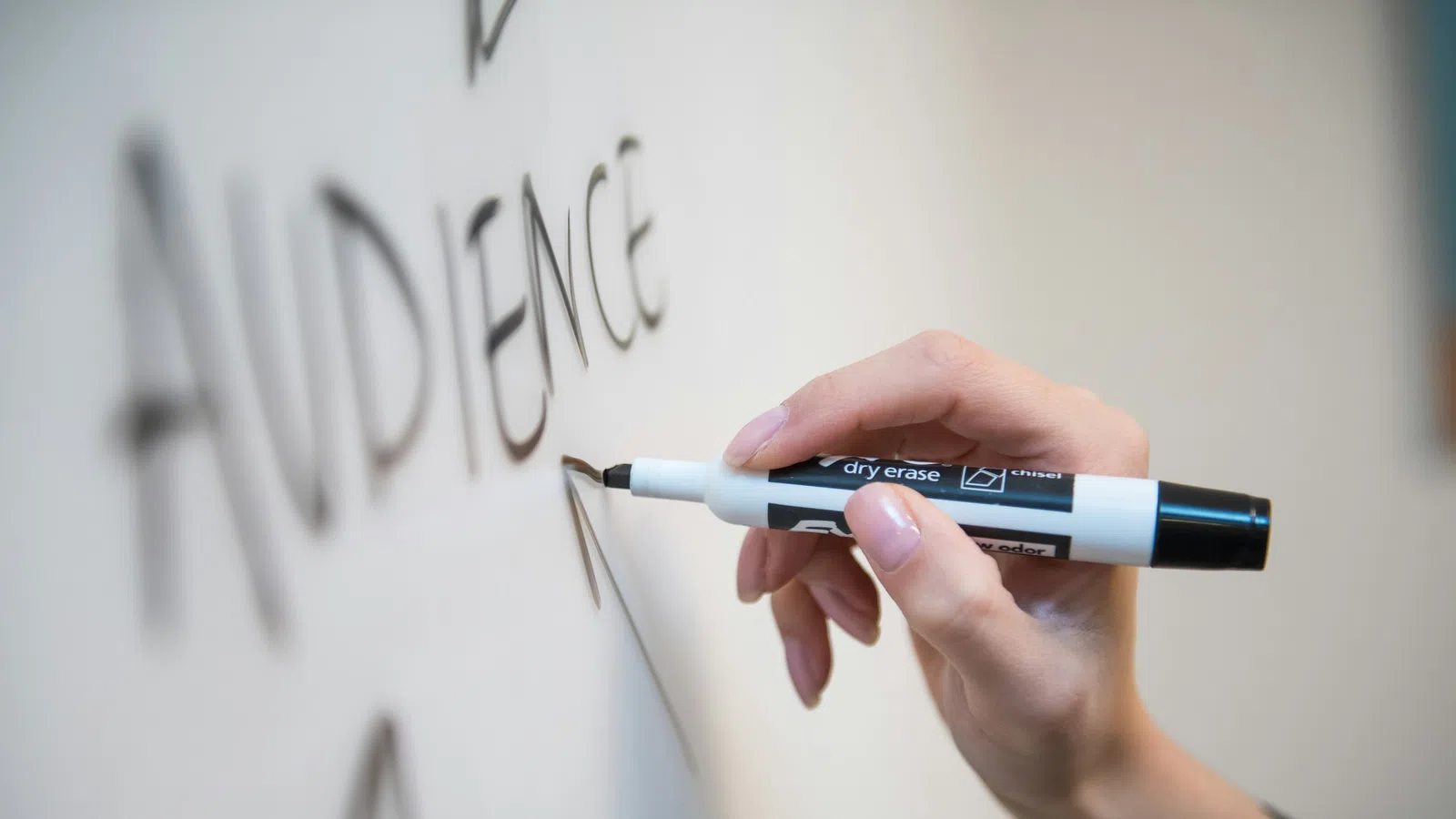 Hand writing the word AUDIENCE in black dry erase marker on a whiteboard.
