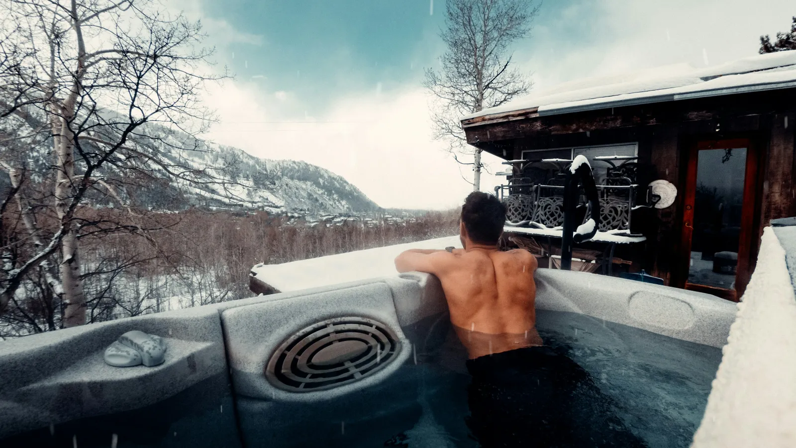 Man relaxing in hot tub on snowy mountain cabin deck with winter landscape and bare trees.