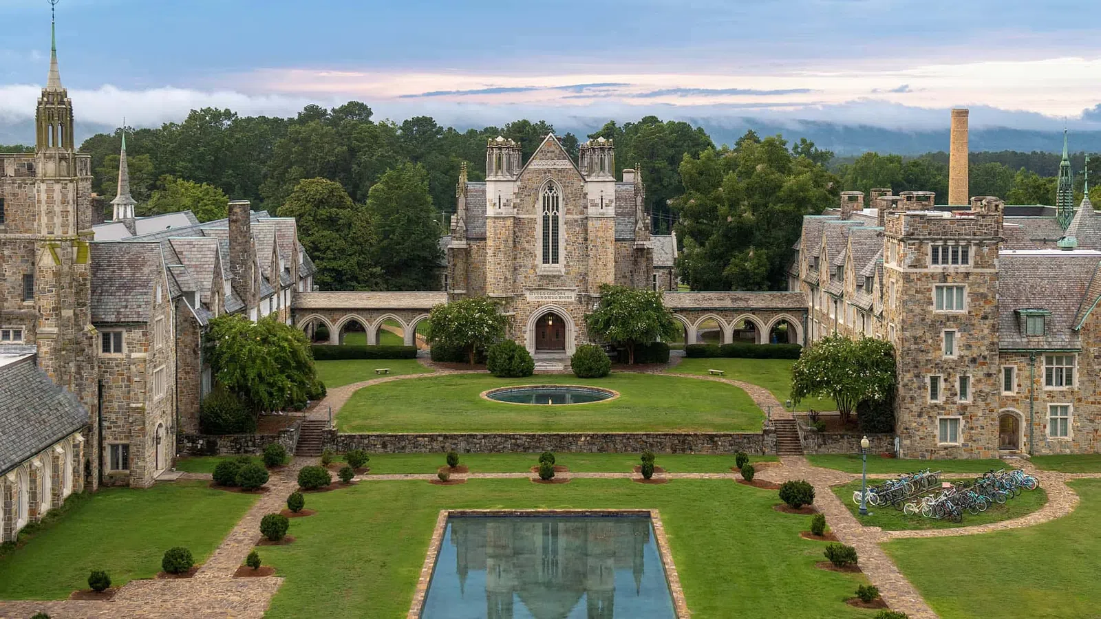 Historic stone college buildings surrounding a manicured lawn with reflective pool and bicycle racks under a cloudy sky