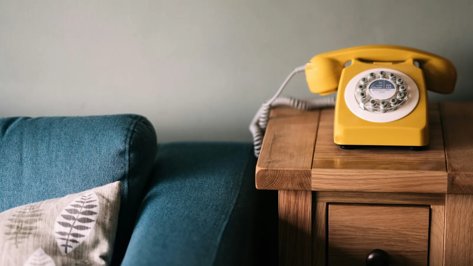Yellow rotary dial phone on wooden side table next to blue couch with patterned pillow