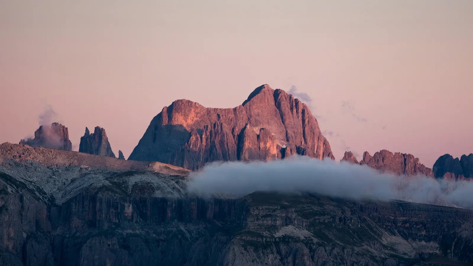 Sunset light casts pink hues on rocky mountain peaks with a layer of low clouds beneath them under a clear sky.