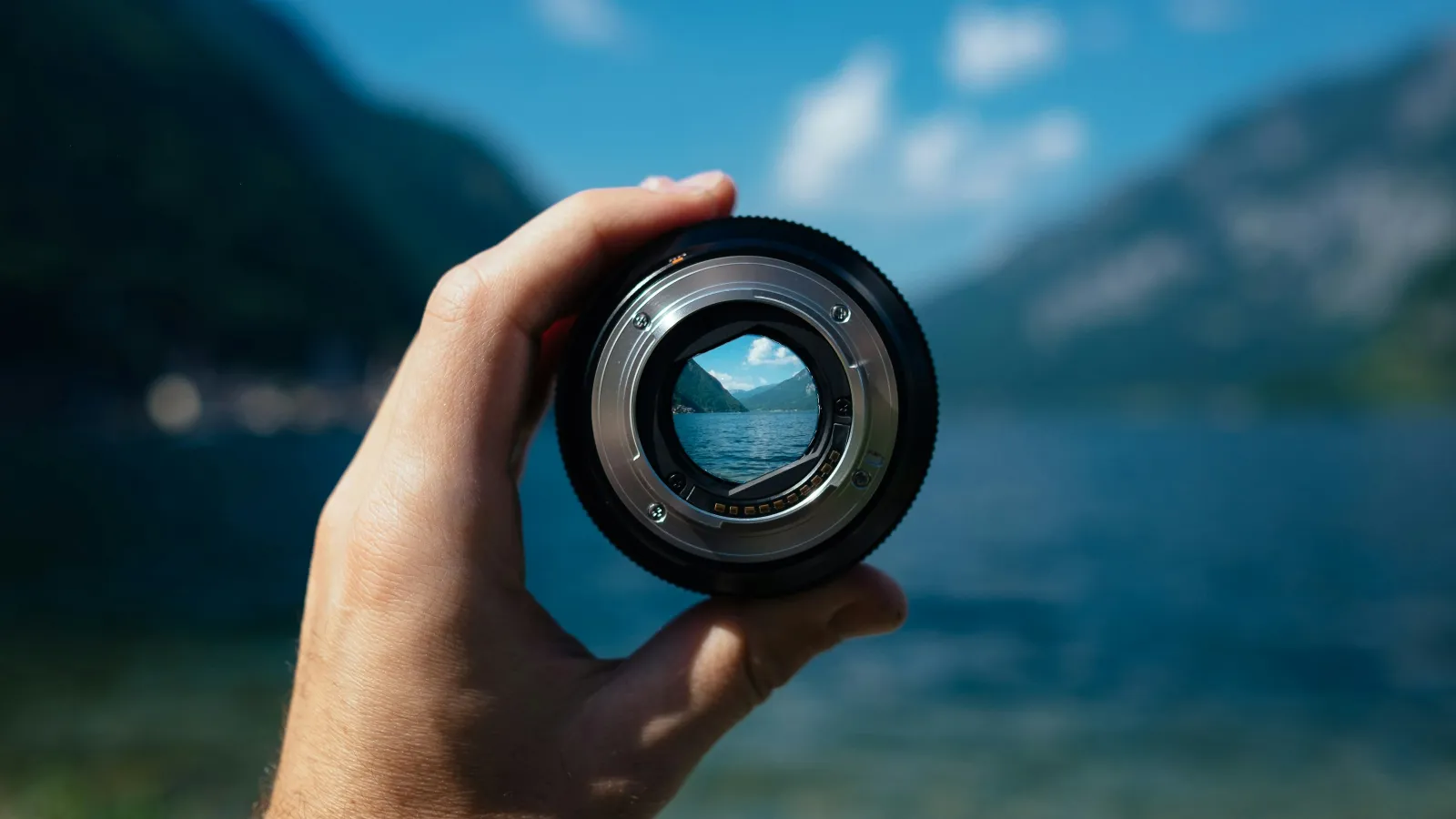 Hand holding a camera lens focusing on a clear lake and mountain scenery in the distance under blue sky.