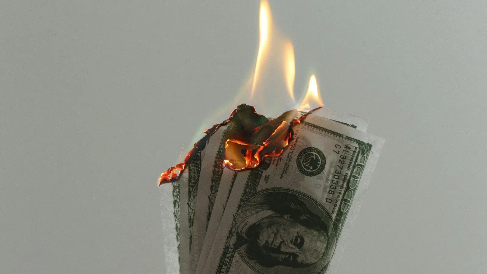Hand holding burning US hundred-dollar bills against a plain background symbolizing financial loss.