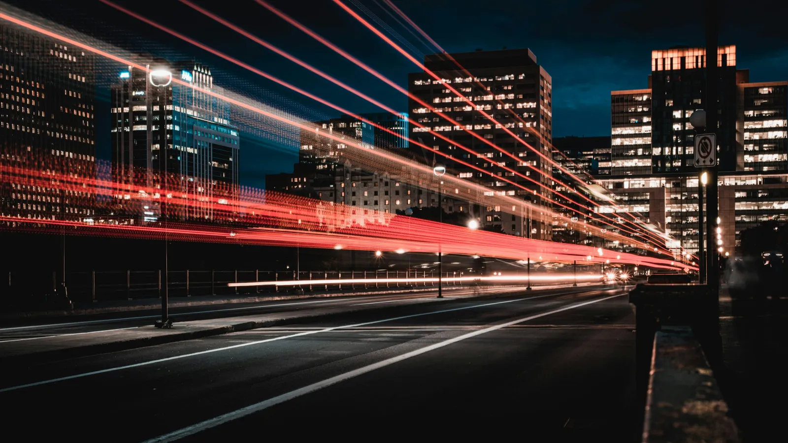City street at night with light trails from moving vehicles against illuminated skyscrapers.