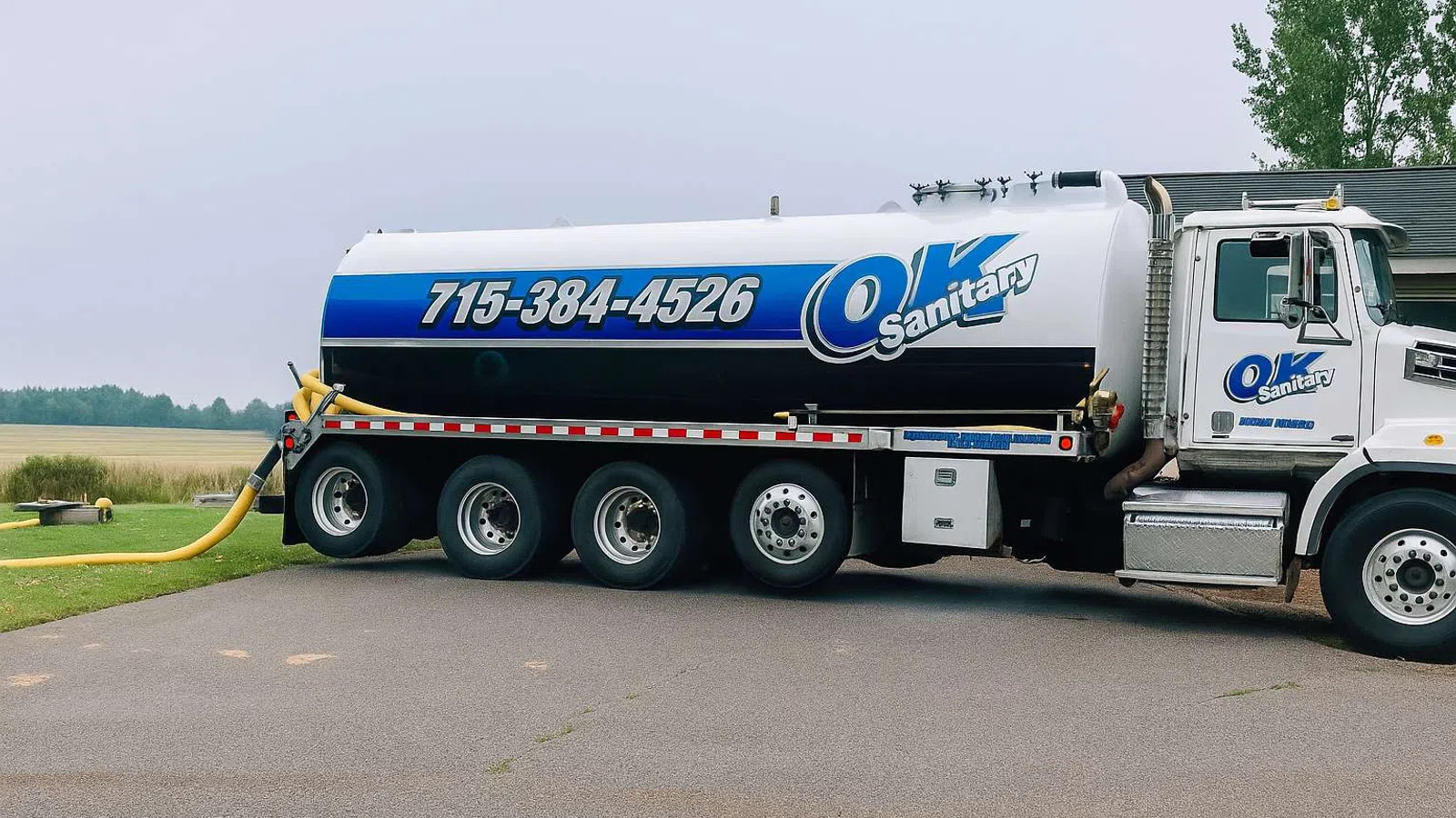 White and blue OK Sanitary tanker truck parked on a driveway with hose extended onto grass.