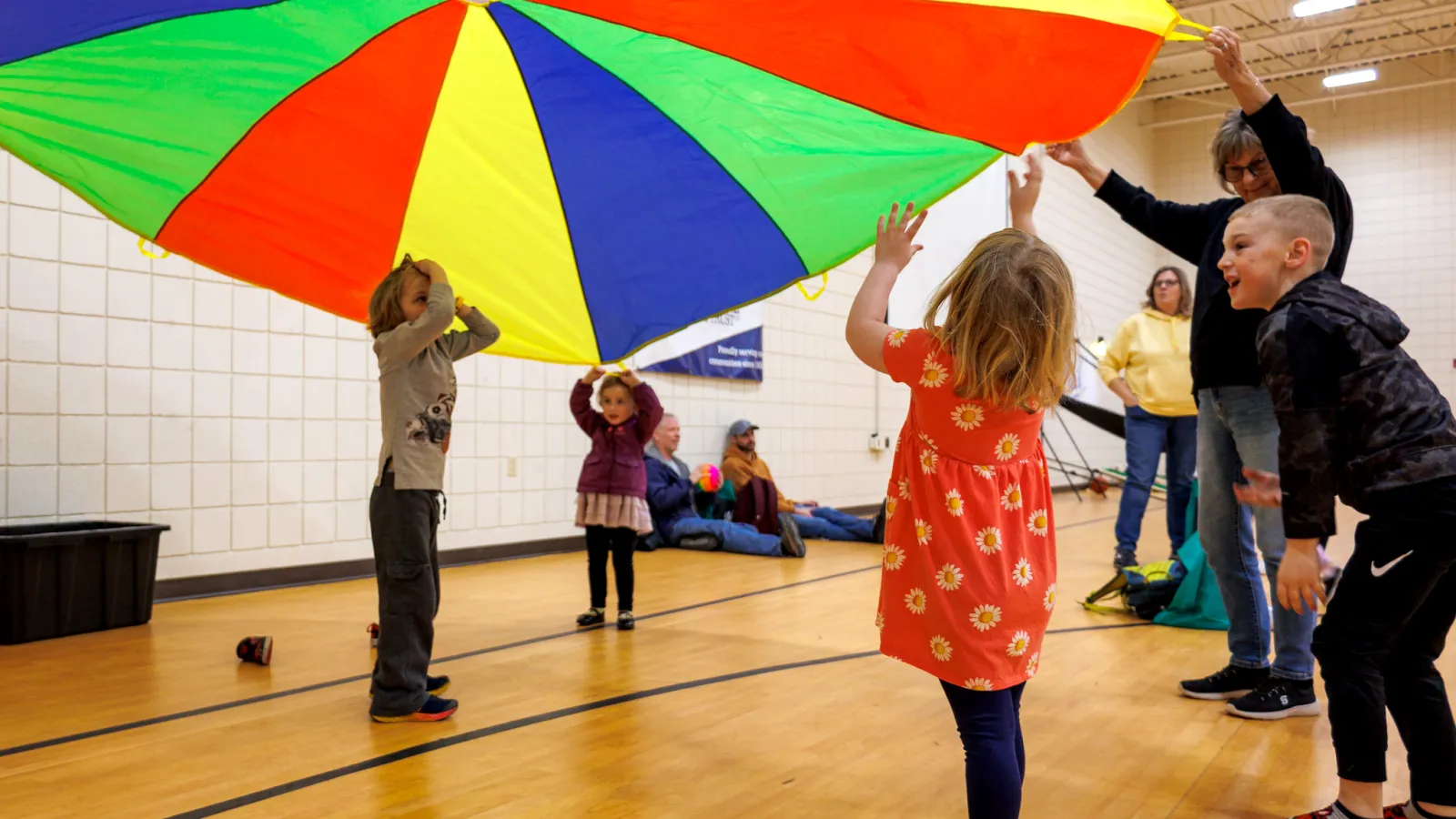 Children and adults playing with a colorful parachute indoors on a gym floor, engaging in a group activity.