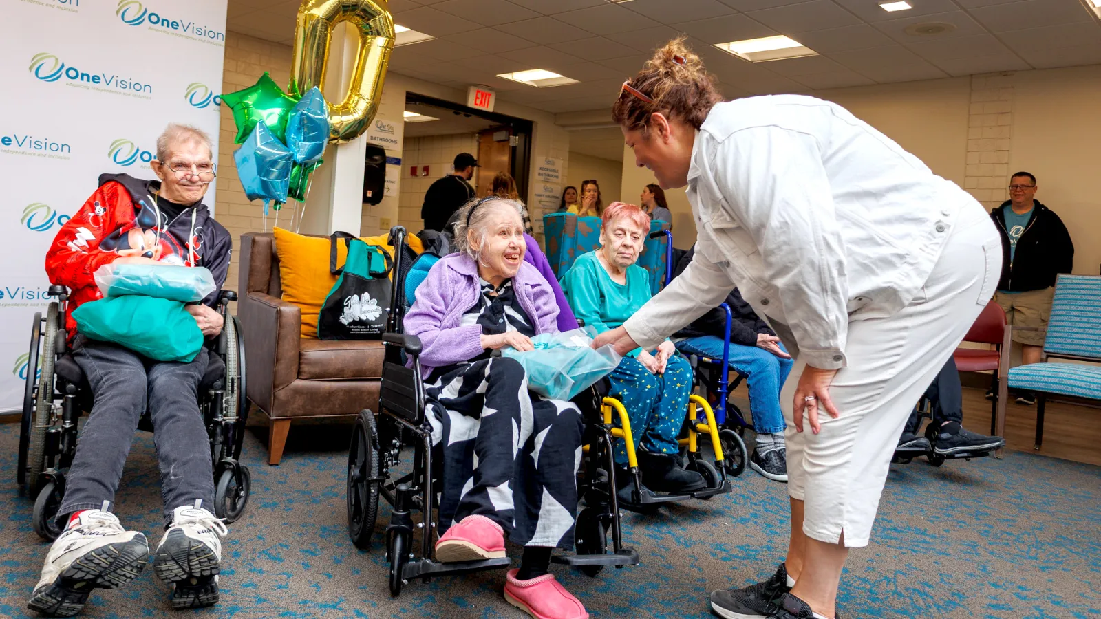 Caregiver gives gift to smiling elderly woman in wheelchair during a celebration at a community center.