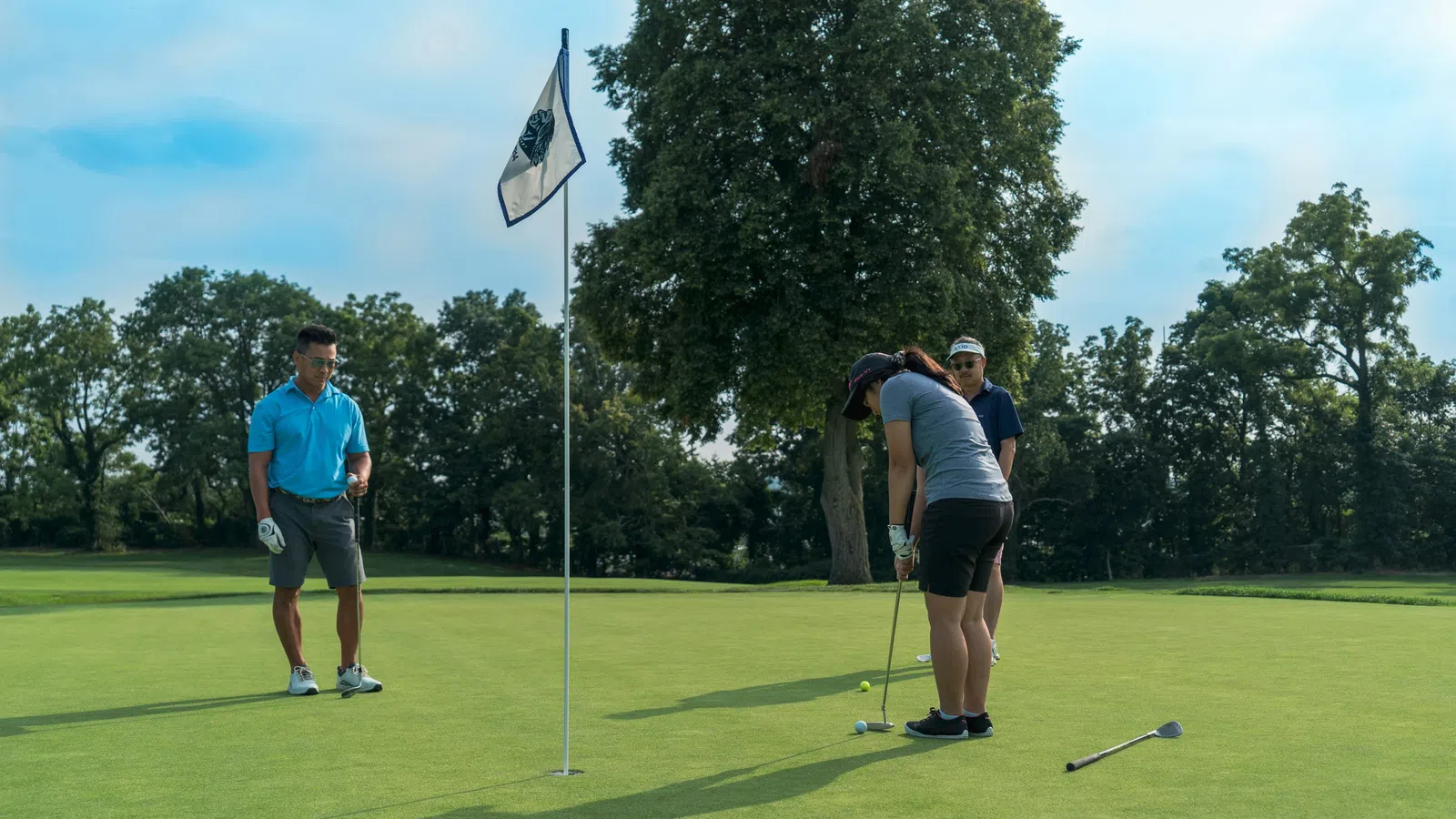 Three golfers on a green putting near a flagstick under a clear sky with trees in the background.