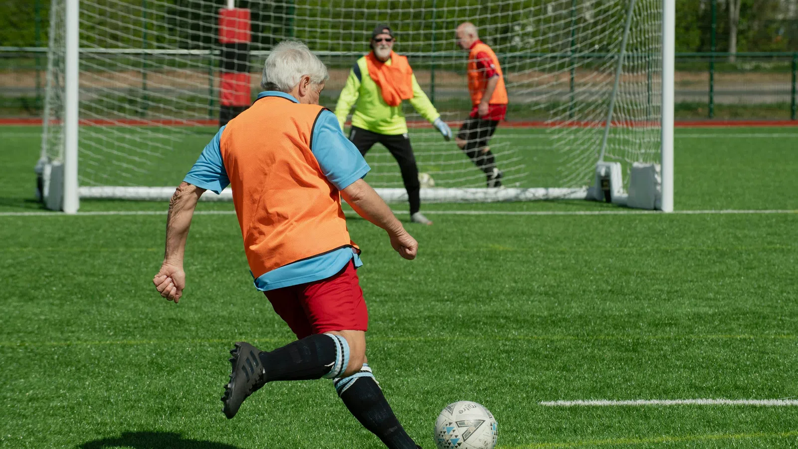 Senior man in orange vest kicking soccer ball toward goal with goalkeeper and defender in background on green field