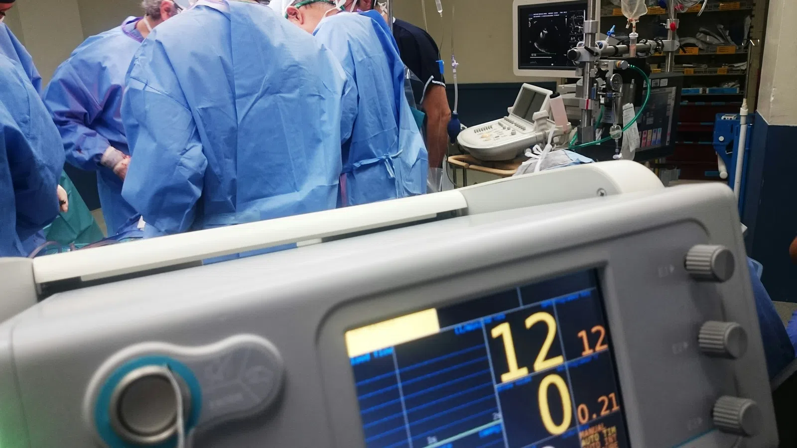 Vital signs monitor in foreground during surgery with medical team in blue scrubs and masks in operating room.