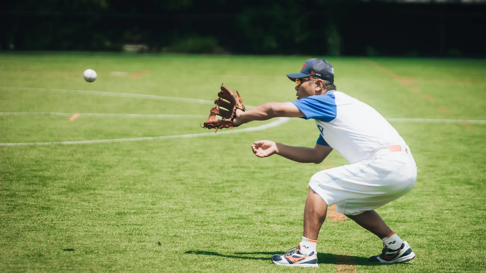 Baseball player in white uniform crouching and catching a ball with a glove on a green field during daytime