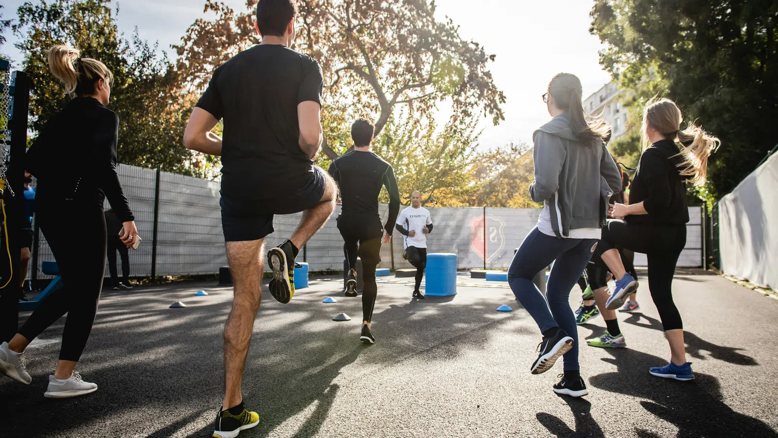 Group of people exercising outdoors in a sunny urban setting, doing leg lifts during a fitness class.