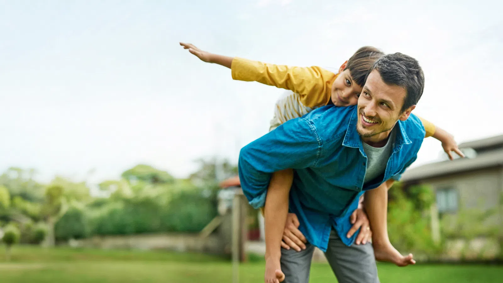 Father giving piggyback ride to son outdoors with greenery and clear sky in background