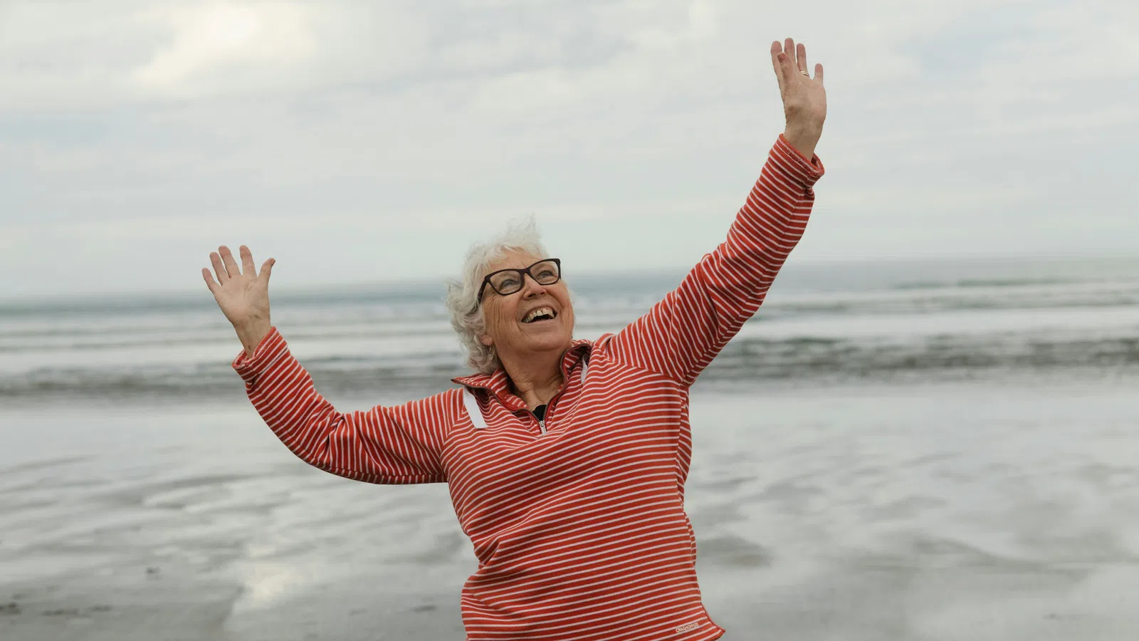 Smiling elderly woman in red striped shirt joyfully raising hands on a cloudy beach with ocean in the background