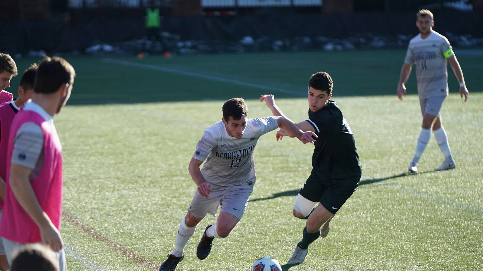 Two soccer players contesting for the ball on a sunlit field with teammates watching from the sidelines.