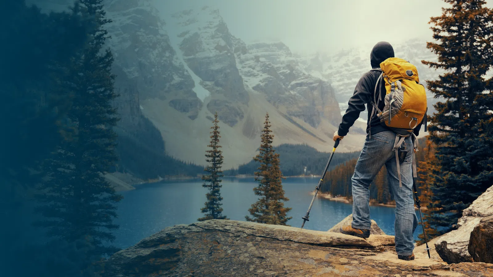 Hiker with yellow backpack overlooks mountain lake surrounded by tall pine trees and rocky peaks.