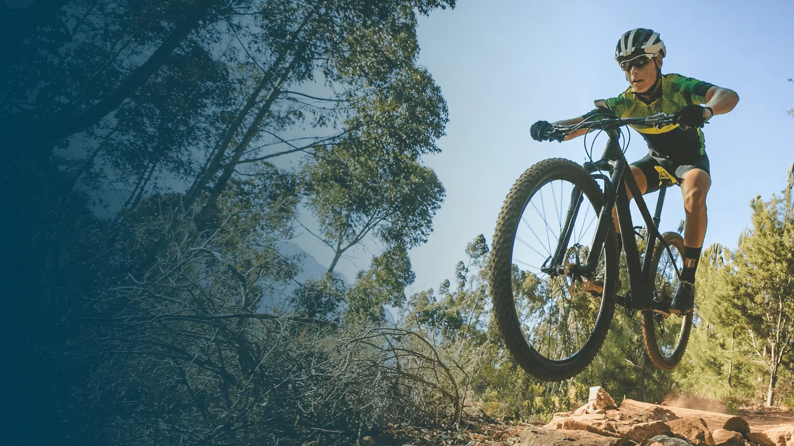 Mountain biker in helmet jumping over rocky trail surrounded by trees under clear blue sky