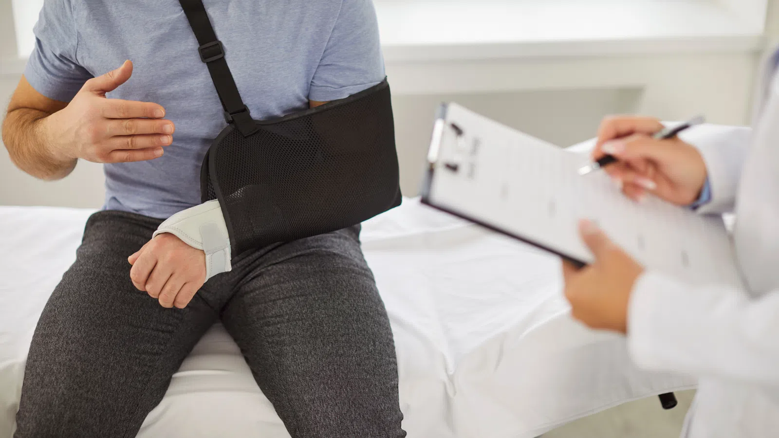 Man with arm in sling consulting doctor taking notes on clipboard in medical office.