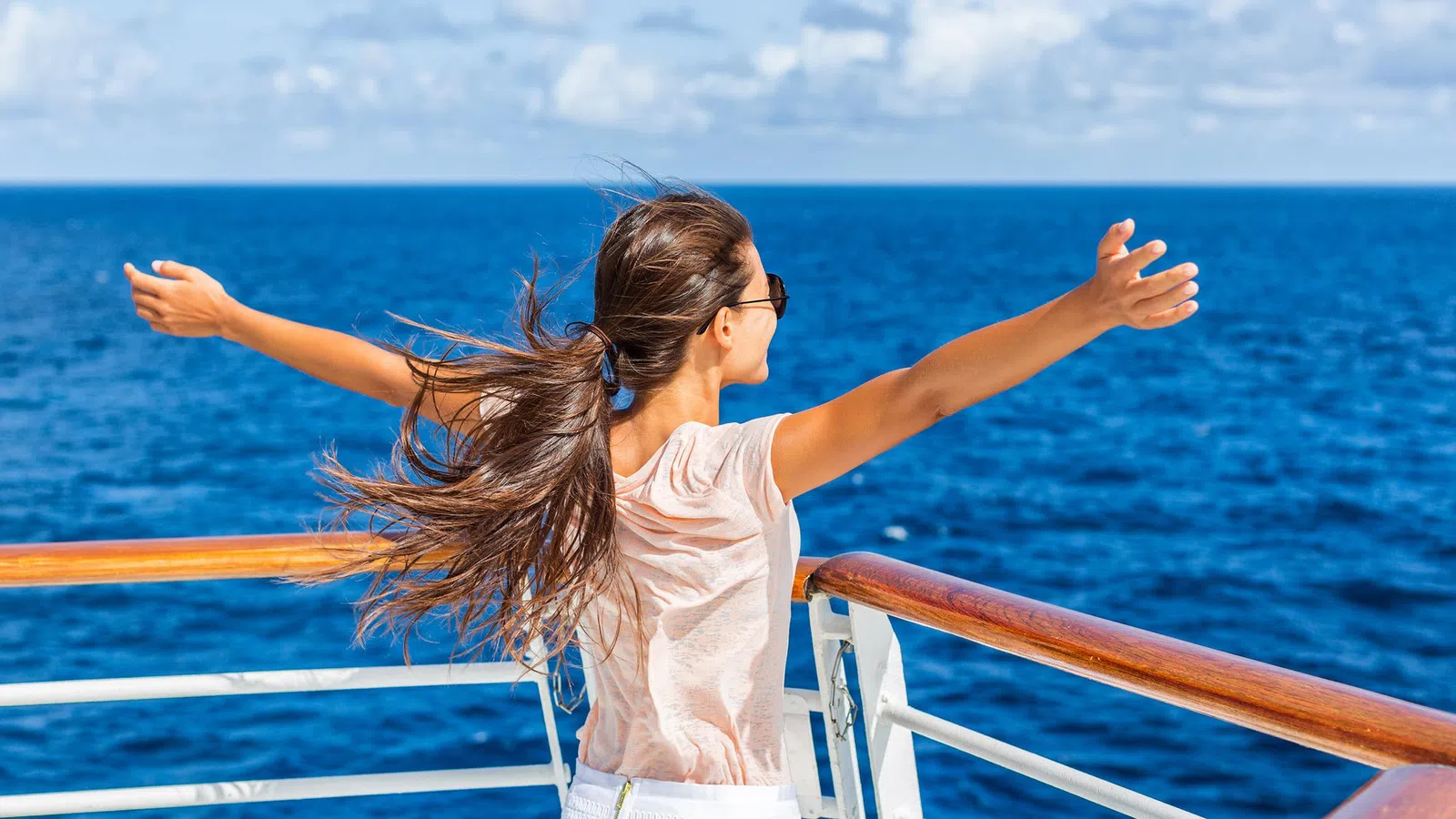 Woman with long hair embraces the ocean breeze on a ship deck under blue sky and clouds.