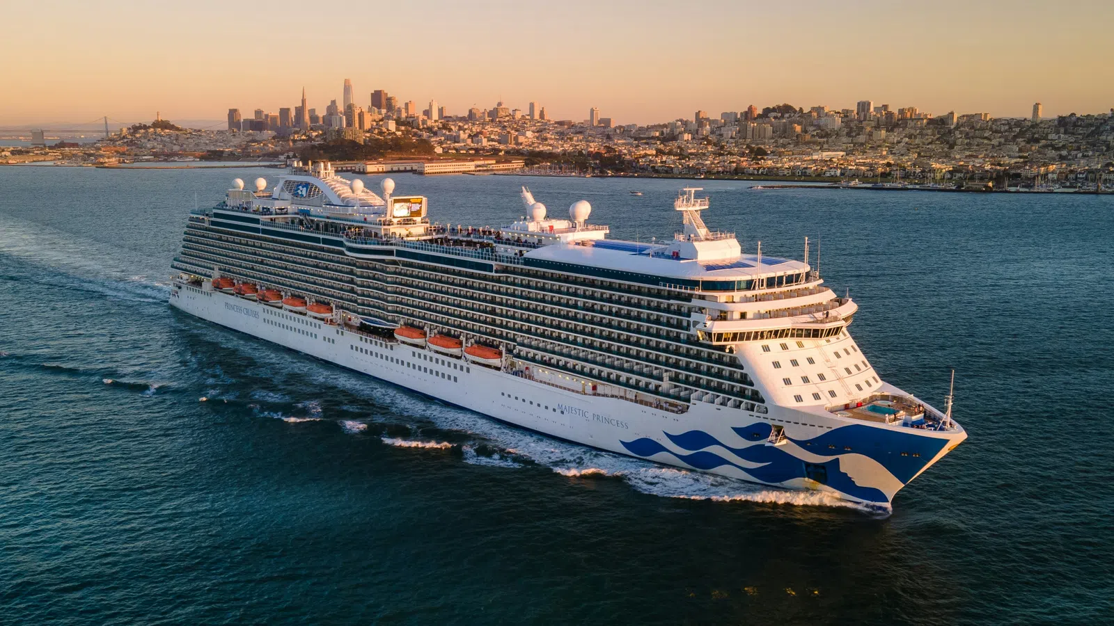 Large cruise ship sailing near a city skyline at sunset with calm blue waters around it