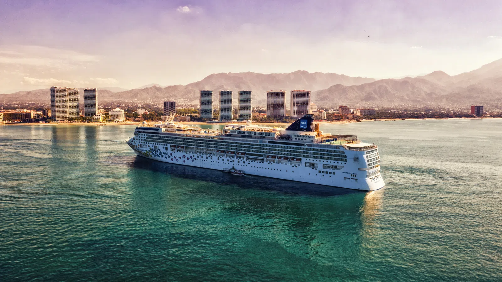 Large cruise ship sailing near a coastal city with high-rise buildings and mountains in the background at sunset.