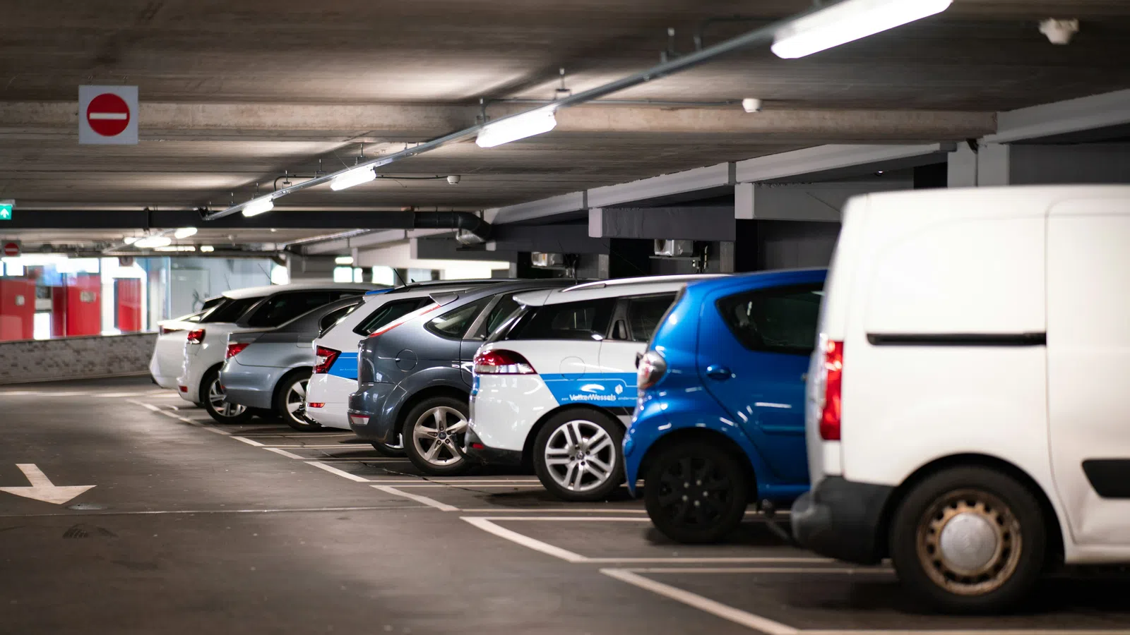 Row of various cars and vans parked in an indoor parking garage with overhead lighting and exit signs.
