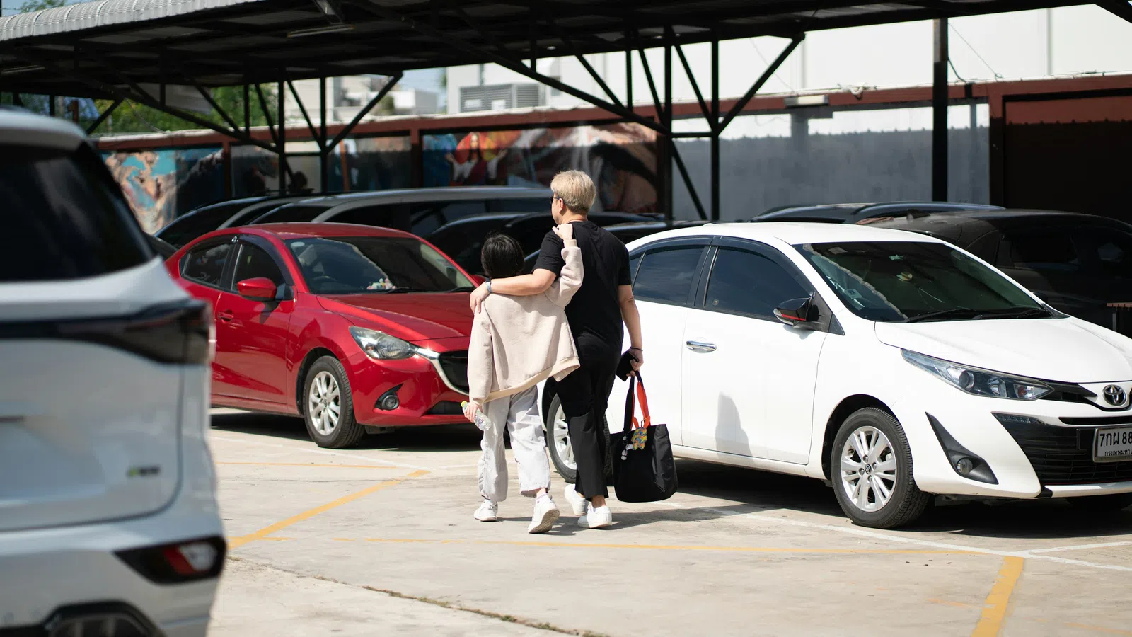 Two people walking arm in arm through a sunlit parking lot with multiple parked cars under a metal canopy.