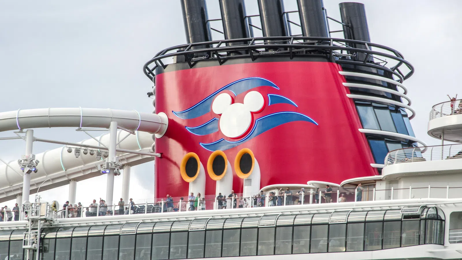Close-up of Disney cruise ship's red funnel with Mickey logo and water slide on upper deck with passengers.