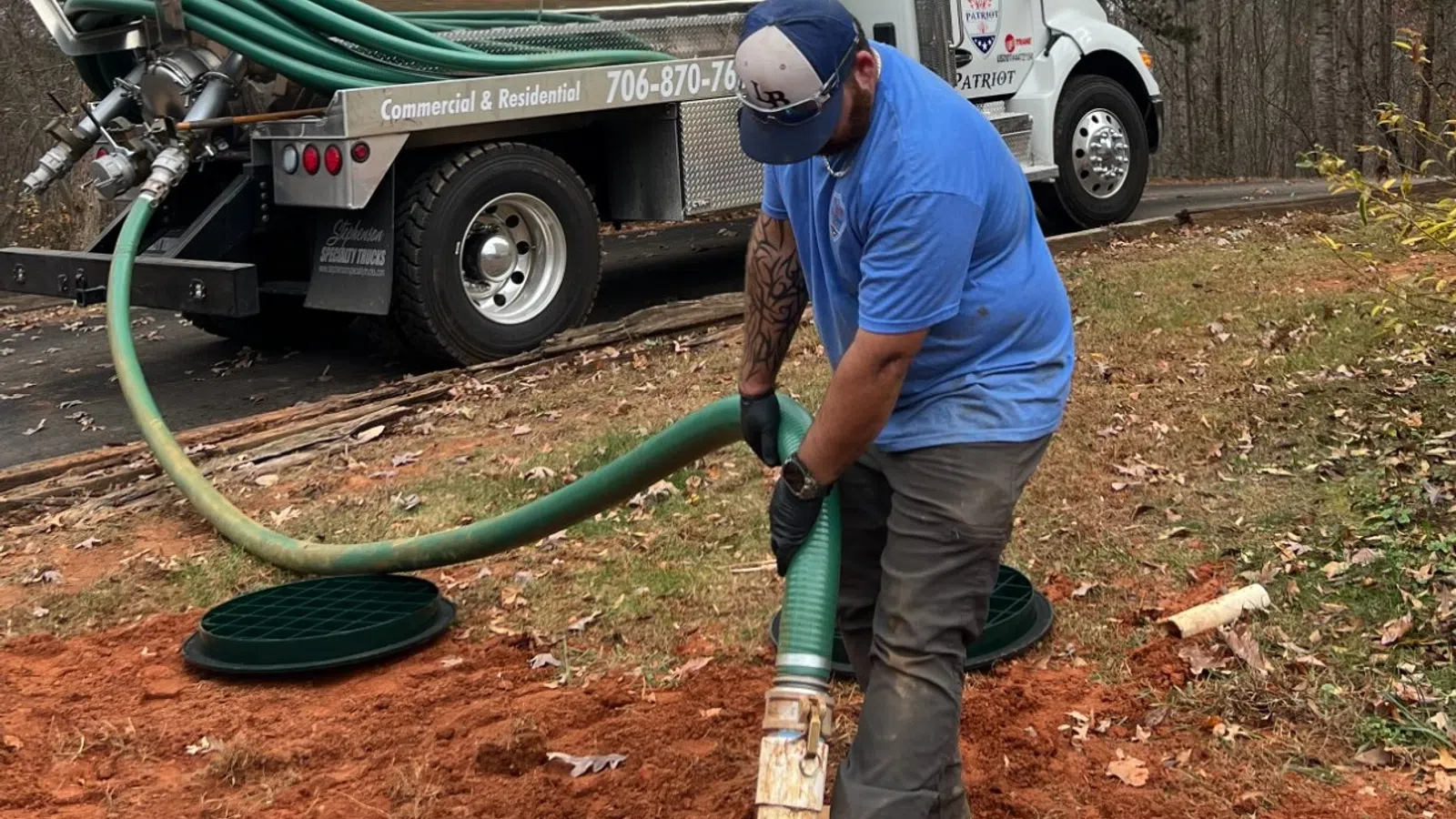 Worker in blue shirt using hose connected to Patriot plumbing truck near open manhole on red dirt ground.
