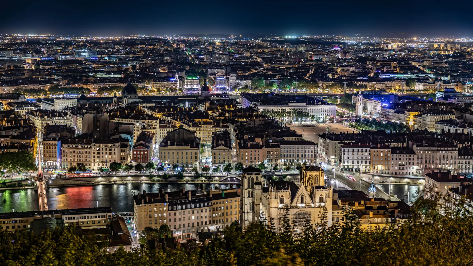 Night view of a European city with illuminated buildings, bridges, river, and streets bustling with lights.
