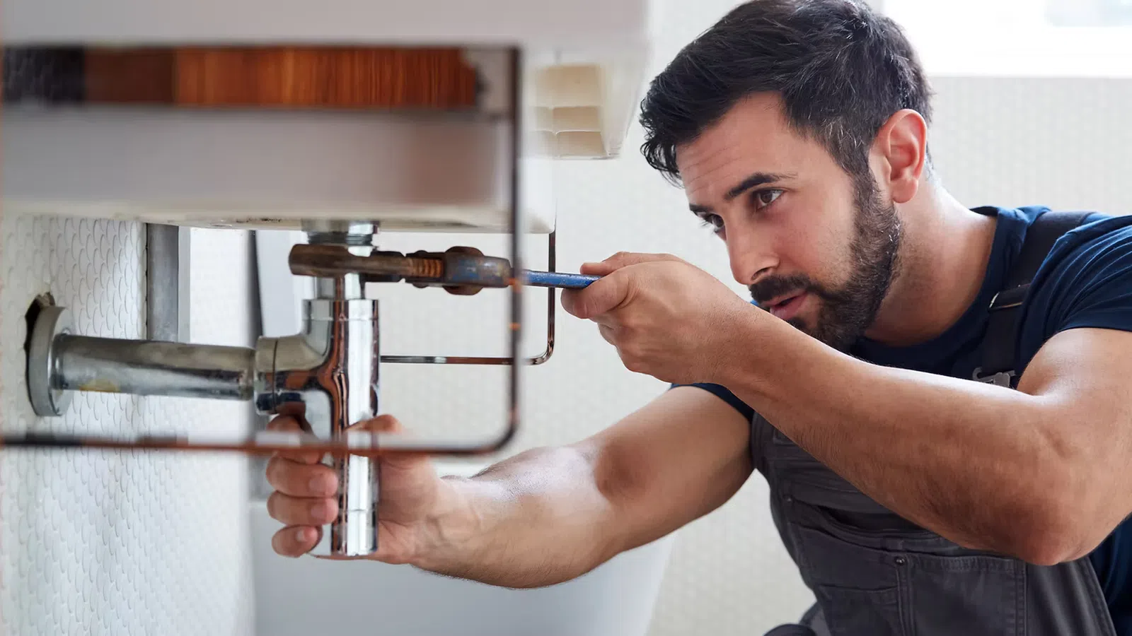 Focused plumber fixing bathroom sink pipes using wrench in a modern bathroom setting.