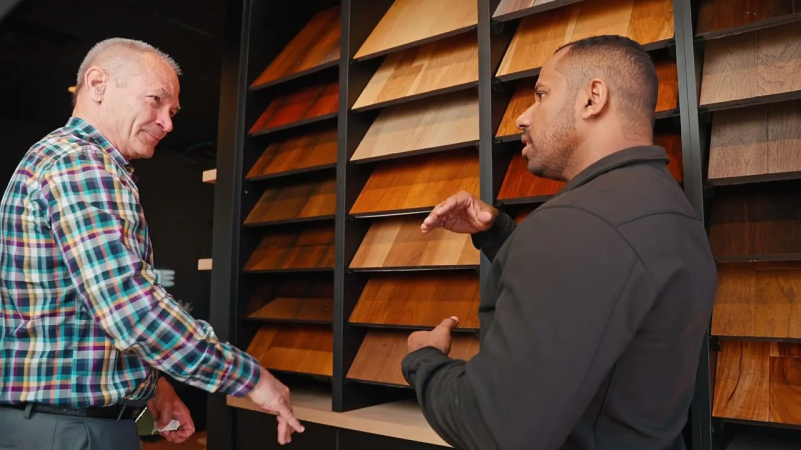 Two men discussing wood flooring samples displayed on a wall in a showroom with various wood finishes.