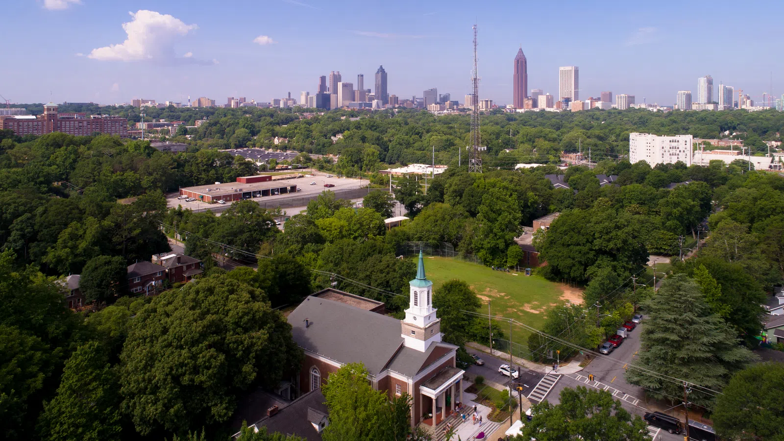 Aerial view of Atlanta skyline with dense green trees and a church with a steeple in the foreground on a sunny day.