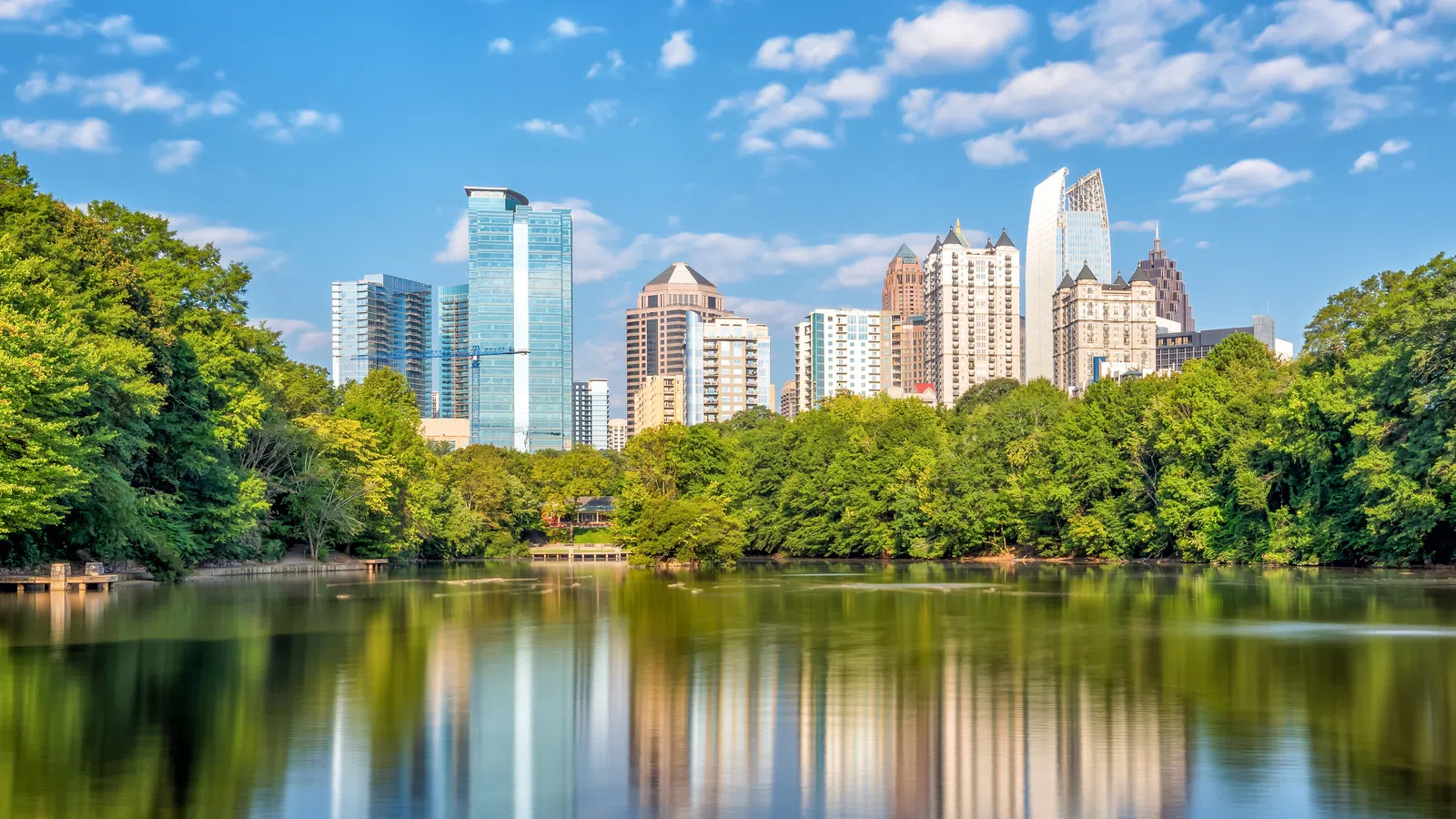 City skyline reflected in calm lake with lush green trees and partly cloudy blue sky above.