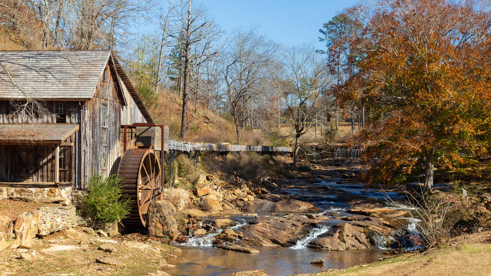 Rustic wooden watermill beside a flowing creek with autumn trees and a clear blue sky in the background
