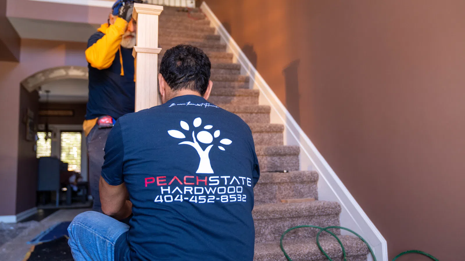 Two workers installing new hardwood stair railing in a home interior with Peach State Hardwood branding visible.