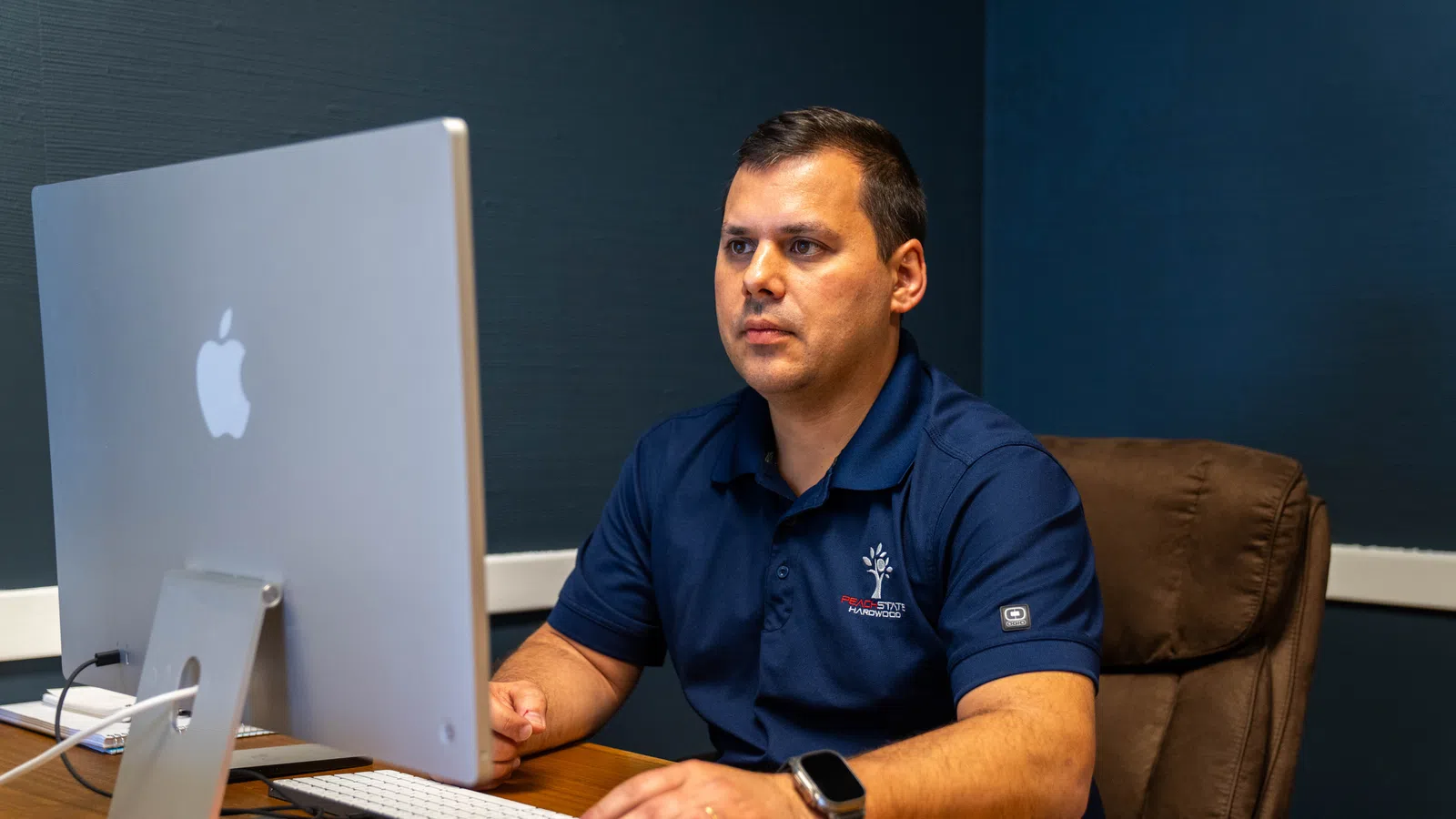 Man in navy blue polo working on an Apple iMac at a wooden desk in a dark office environment