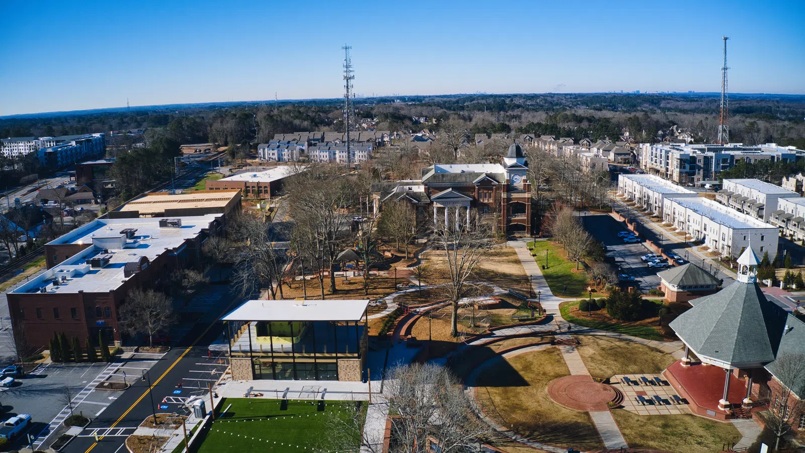 Aerial view of a town park with pathways, pavilion, green spaces, and surrounding buildings under clear blue sky.