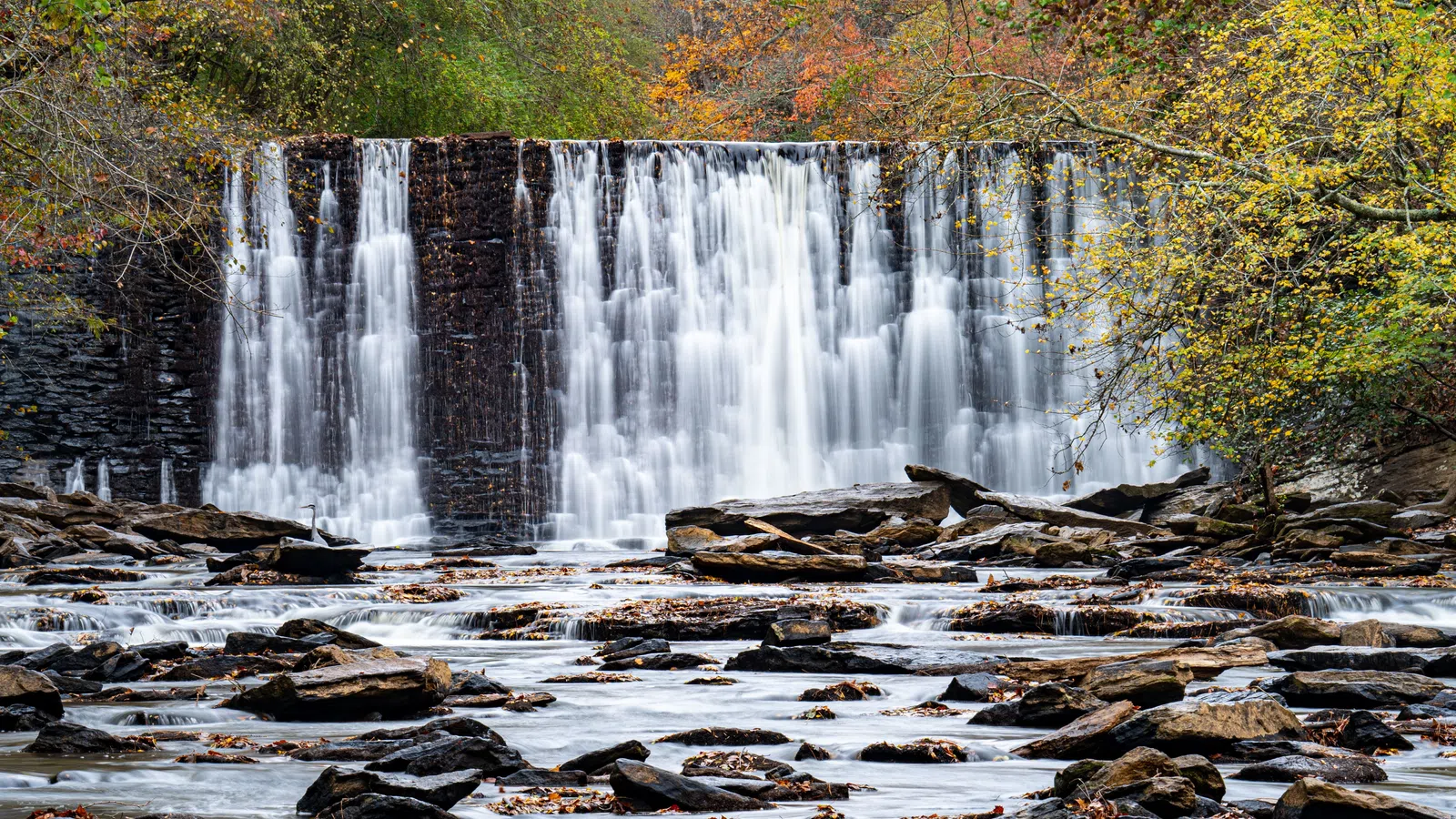Wide waterfall cascading over rocky ledge surrounded by autumn trees with colorful fall foliage.